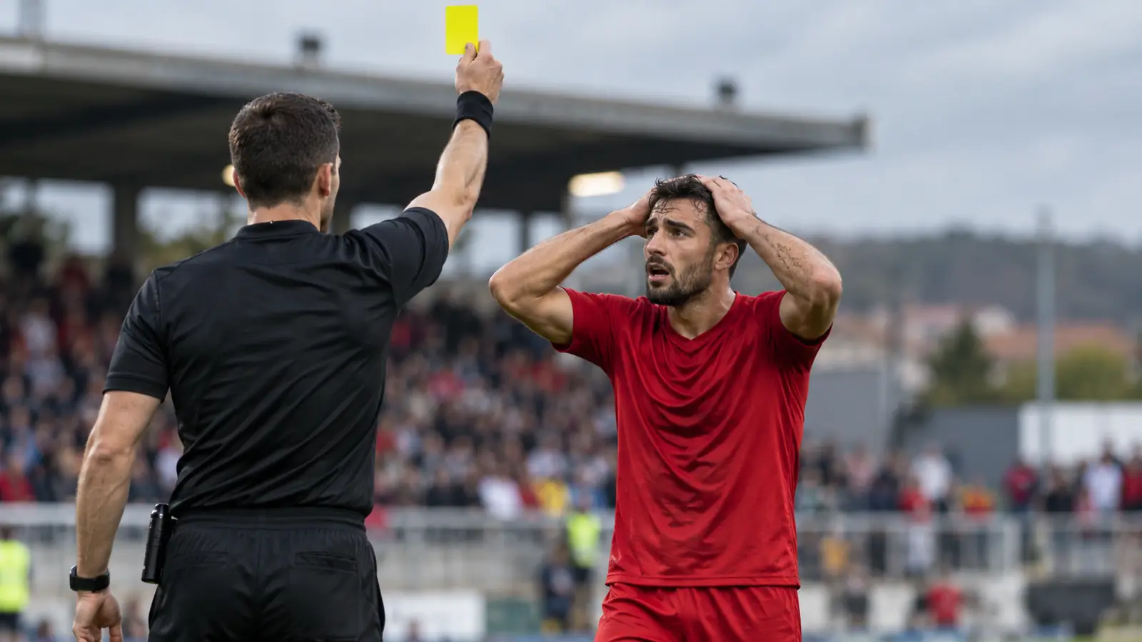 Árbitro muestra una tarjeta amarilla a un defensa con camiseta roja que se lleva las manos a la cabeza en un partido de Segunda