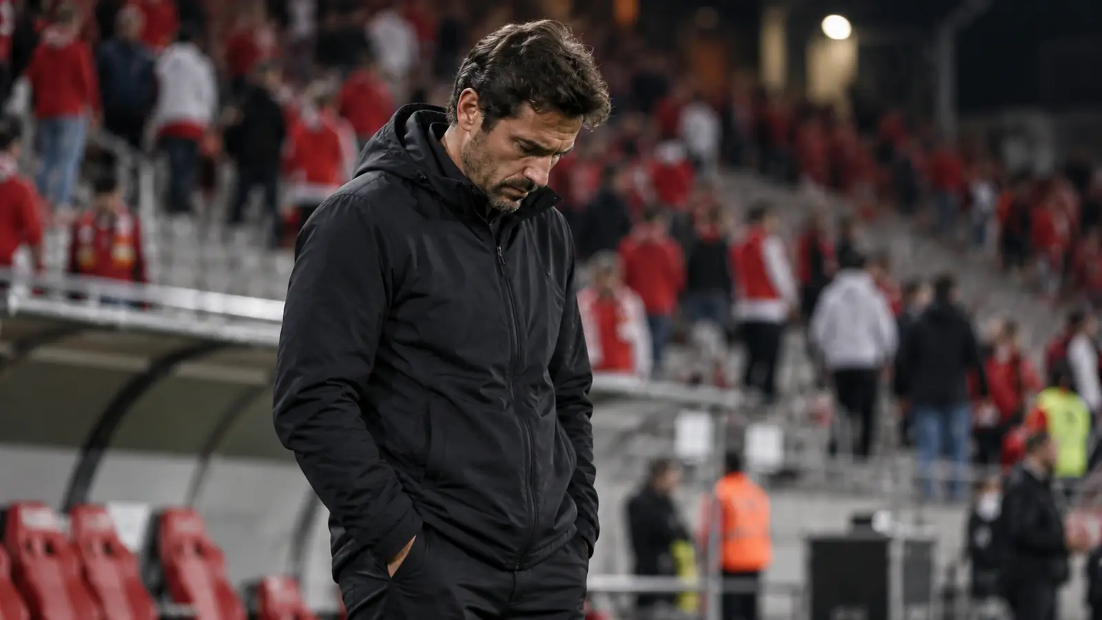 Entrenador cabizbajo en la zona técnica Entrenador cabizbajo en la zona técnica de un estadio al anochecer mientras los aficionados de rojo abandonan la grada