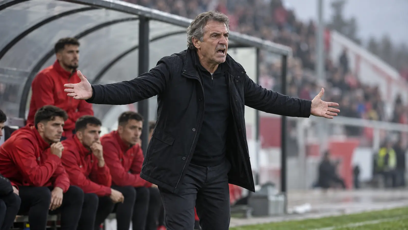 Entrenador protesta con los brazos abiertos en la banda mientras los suplentes con chaquetas rojas miran desde el banquillo