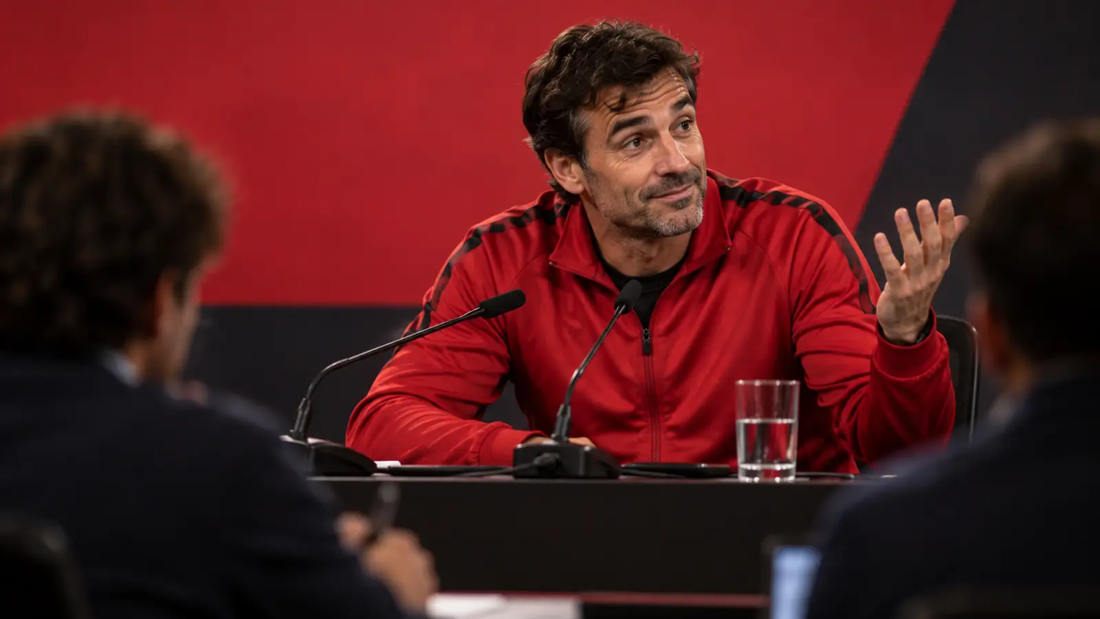 Entrenador en rueda de prensa respondiendo con ironía Entrenador con chaqueta roja gesticula con media sonrisa ante los micrófonos en una rueda de prensa previa al partido