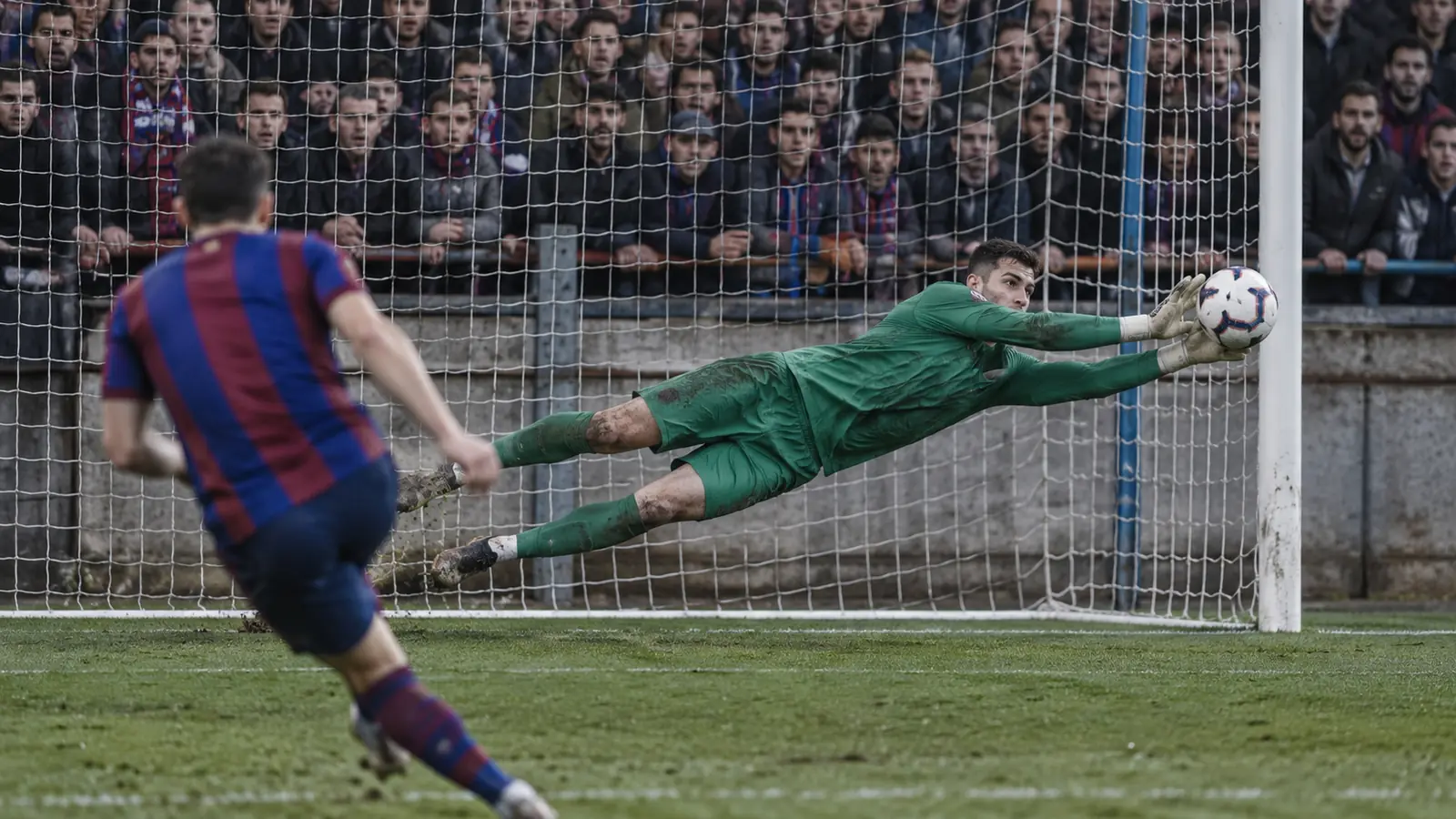 Portero con equipación verde se estira y detiene un penalti junto al poste ante una grada pegada al campo, como la parada de Arnau Tenas en el Eibar-Mallorca